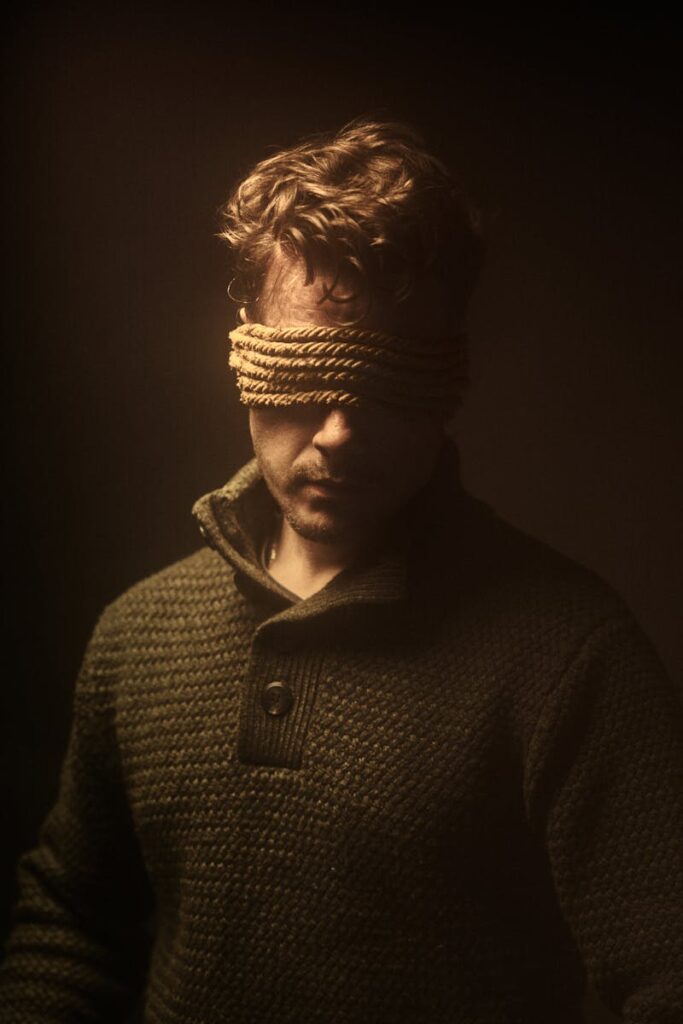 Moody artistic portrait of a blindfolded man in a sweater against a dark background.